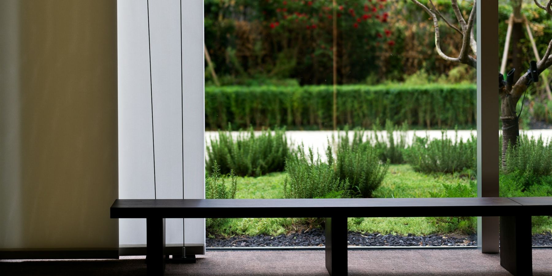 A single low timber bench in front of a floor-to-ceiling window onto a garden.
